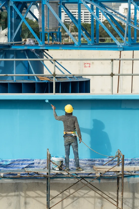 Chantier de construction extérieur avec ouvrier en casque jaune appliquant peinture bleue sur façade depuis échafaudage