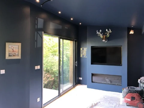 Intérieur moderne en cours de finition avec murs bleus foncés, grandes baies vitrées coulissantes donnant sur jardin, télévision murale et cheminée encastrée contemporaine