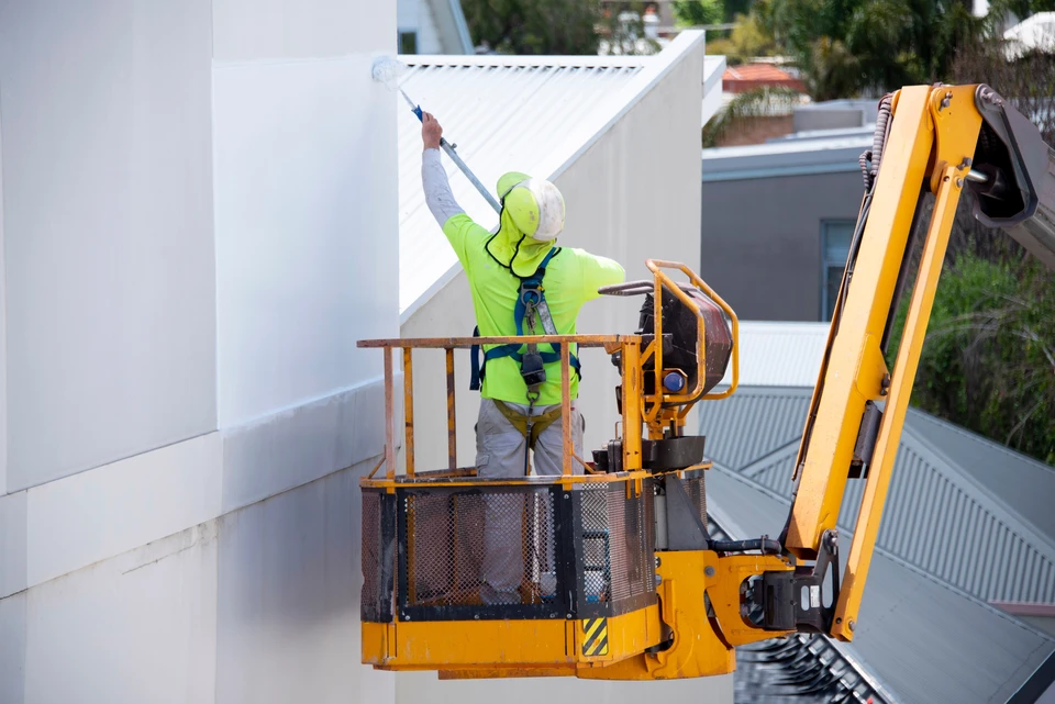 Ouvrier en combinaison bleue et harnais de sécurité rouge travaillant sur nacelle élévatrice jaune devant immeuble de bureaux moderne avec façade vitrée